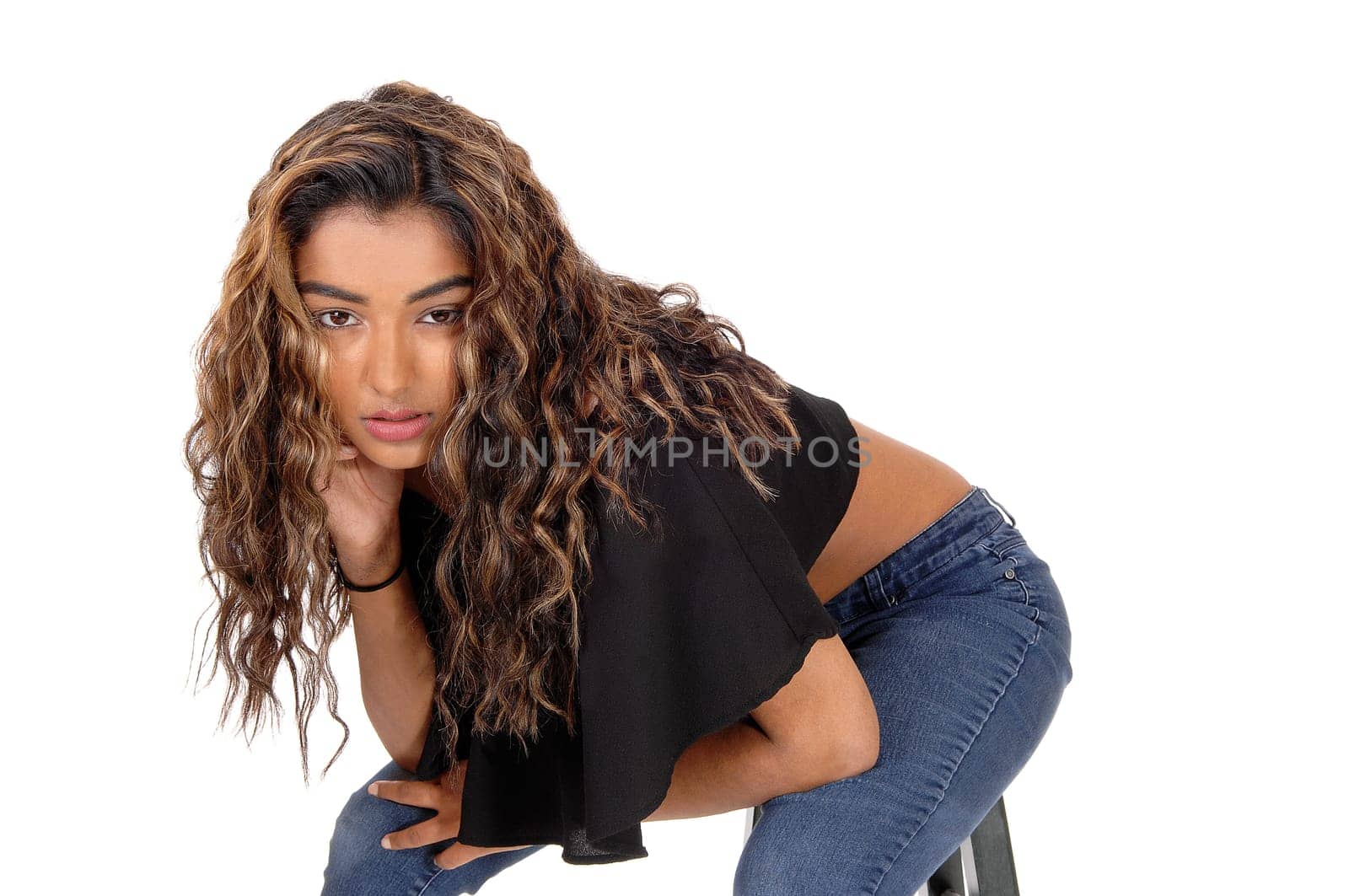 A serious looking young woman sitting on a chair and bending forward.with her long brunette curly hair, isolated for white background.