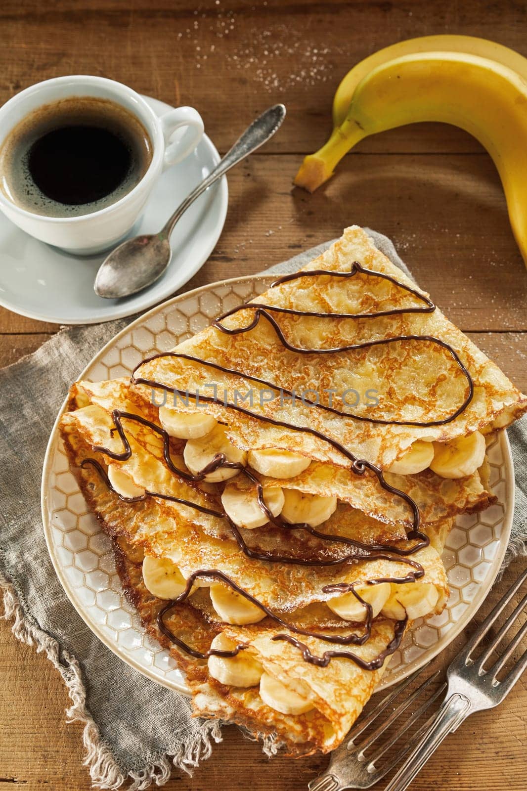 Golden fried batter crepes with banana filling dusted with sugar and drizzled with chocolate viewed top down served with espresso coffee on a rustic table