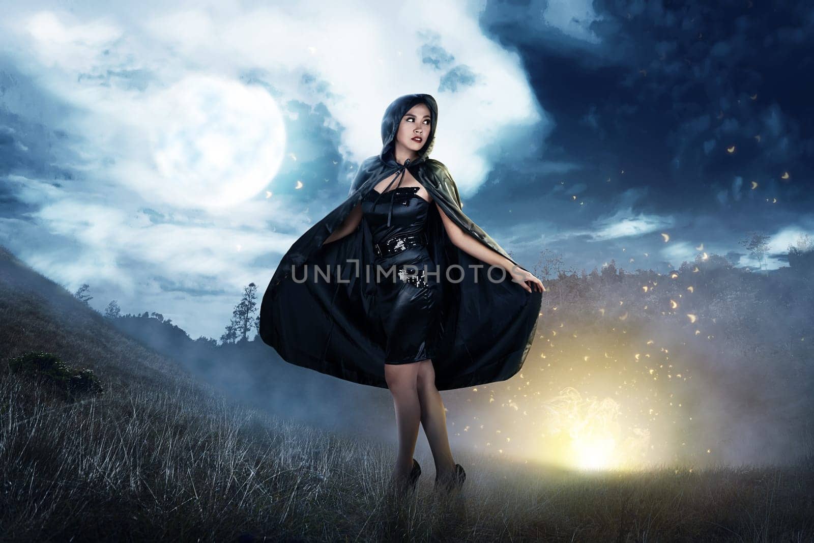 Smiling asian witch woman with black hooded cloak standing at night