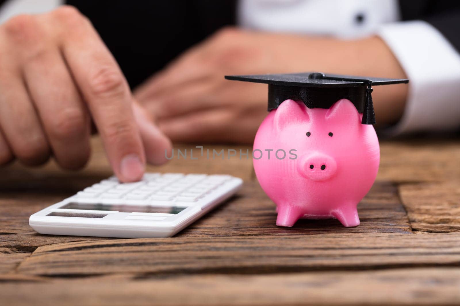 Businessperson's hand using calculator beside piggybank with graduation hat