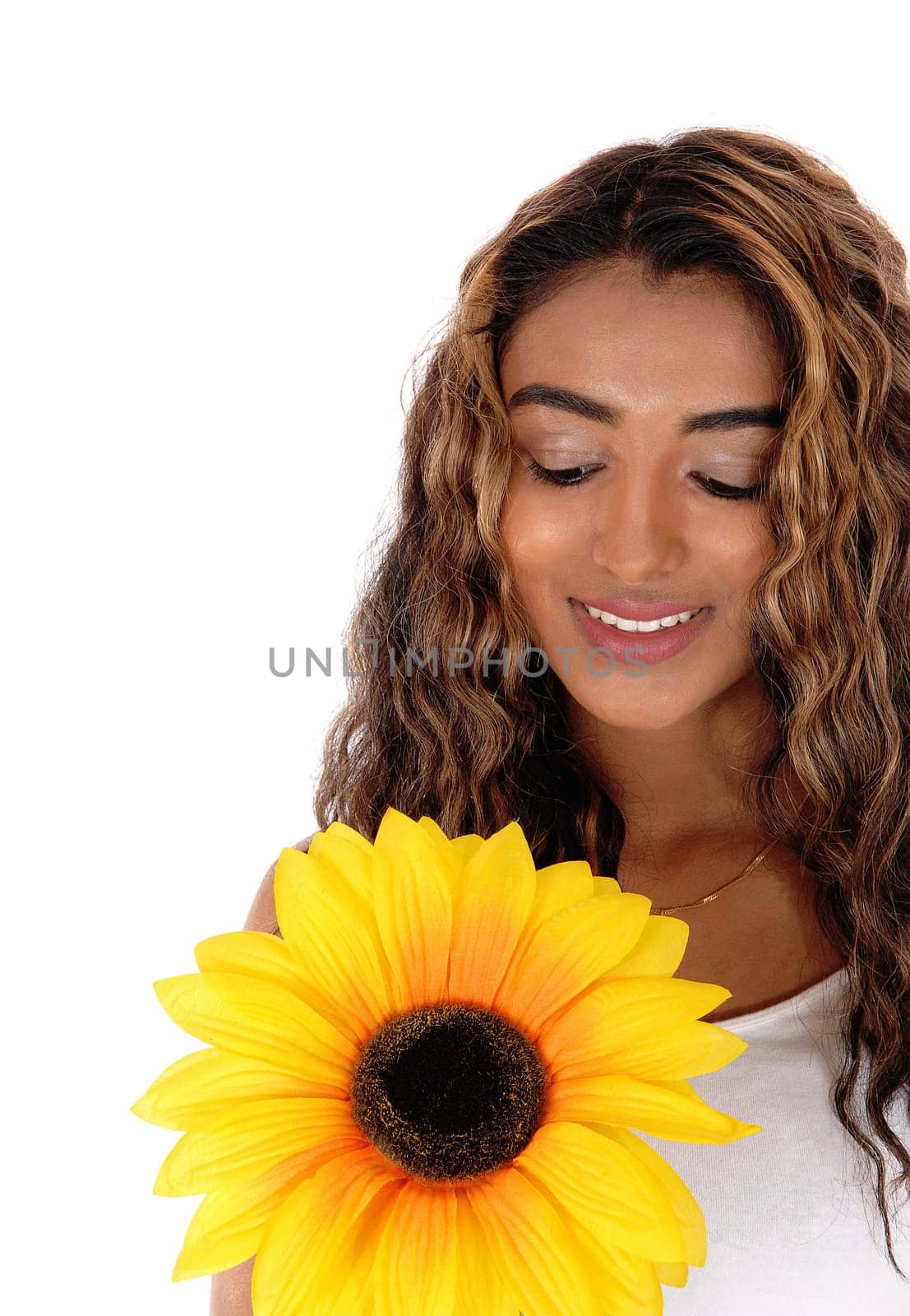 A happy smiling teen girl with long curly brunette hair looking at the .sunflower she holds in her hand, isolated for white background.