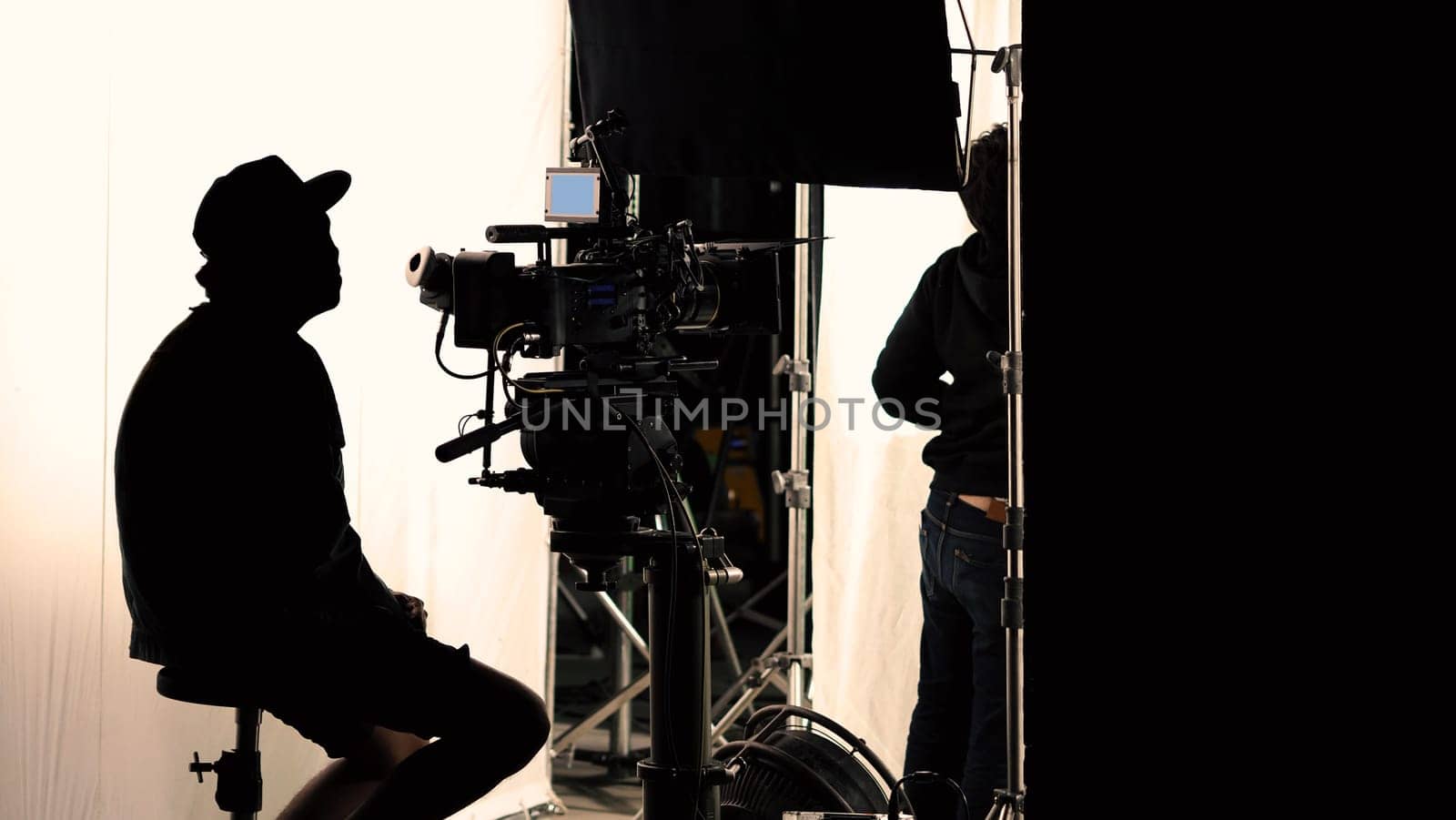 Video production behind the scenes which film crew team in silhouette shooting or recording tv movie commercial with professional equipment such as high definition 4k camera with monitor and tripod or gimbal rig in big studio set.