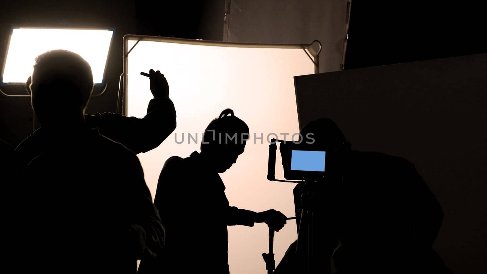 Video production behind the scenes which film crew team in silhouette shooting or recording tv movie commercial with professional equipment such as high definition 4k camera with monitor and tripod or gimbal rig in big studio set.