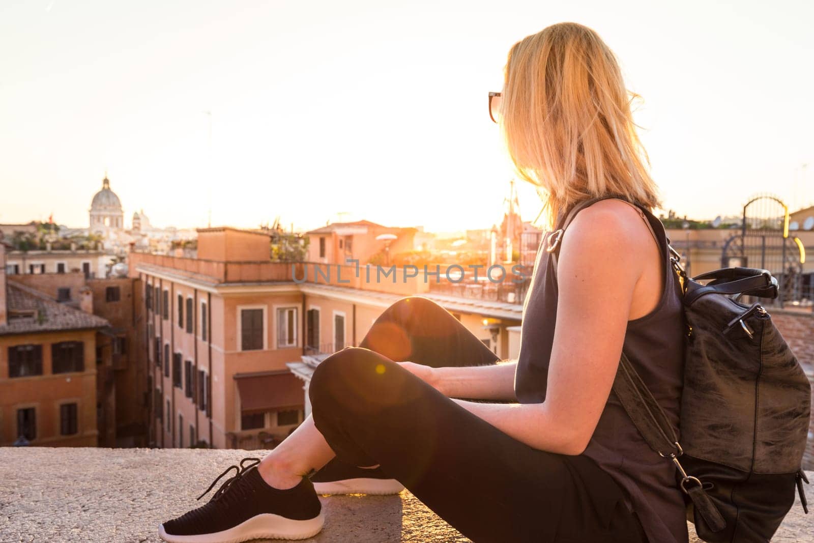 Fashinable female tourist with vintage hipster backpack admiring the beautiful view of at Piazza di Spagna, landmark square with Spanish steps in Rome, Italy at sunset.