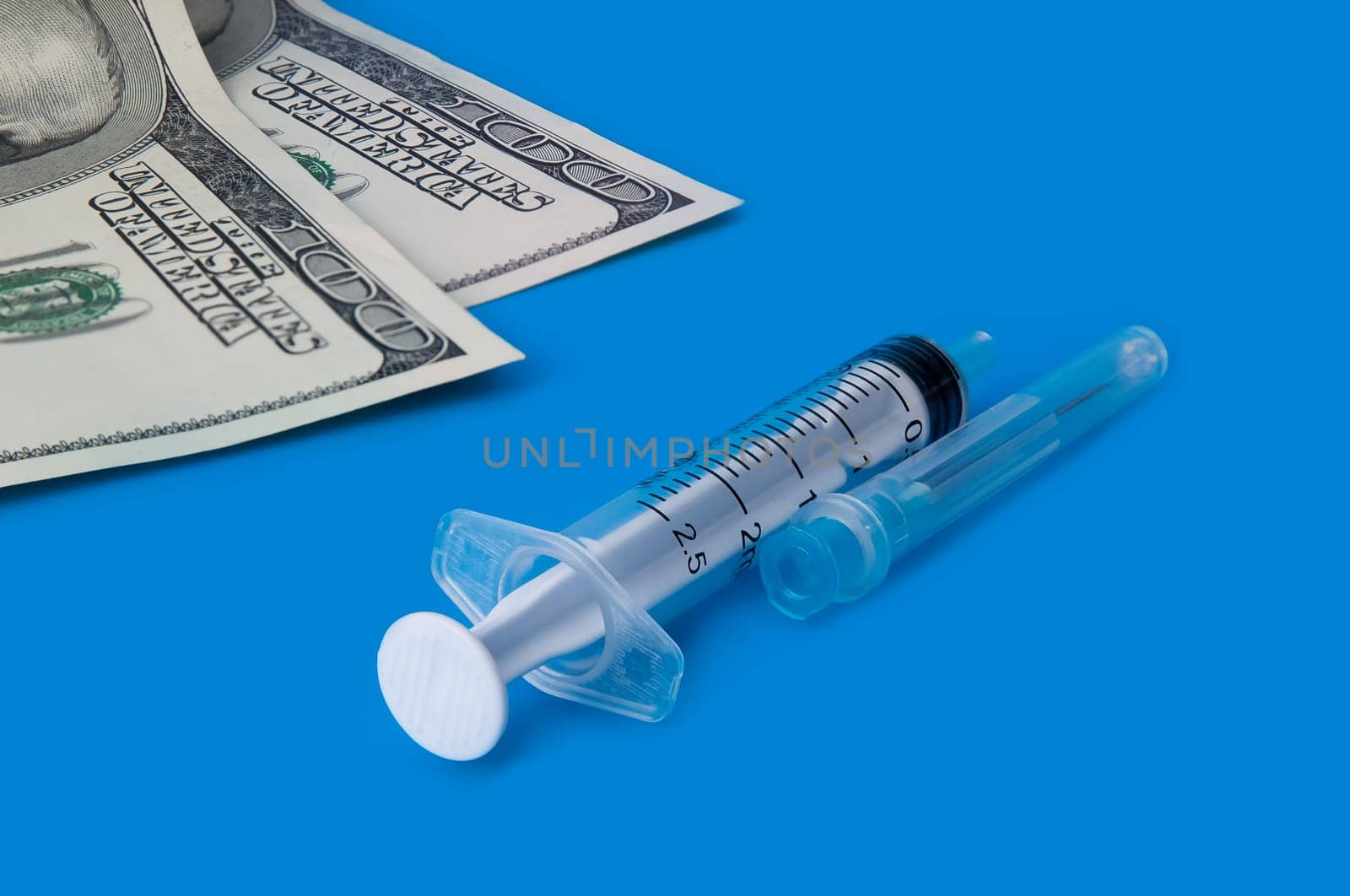 A syringe, and one hundred dollar bills on a blue background. Side view.
