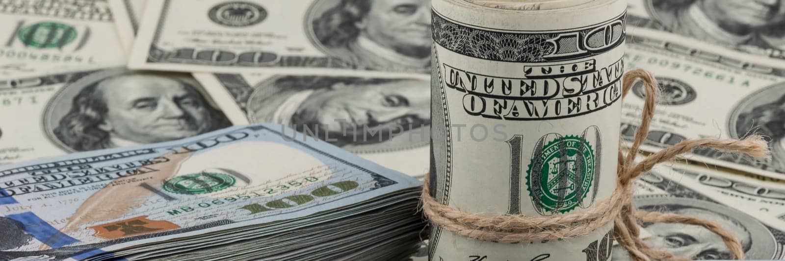 A roll of one hundred dollar bills tied with a rope against the backdrop of scattered dollars.