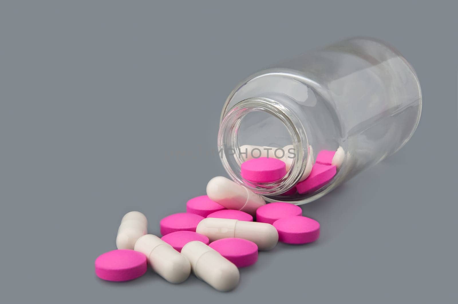 White pills and pink tablets are scattered from a glass bottle on a gray background. Side view.