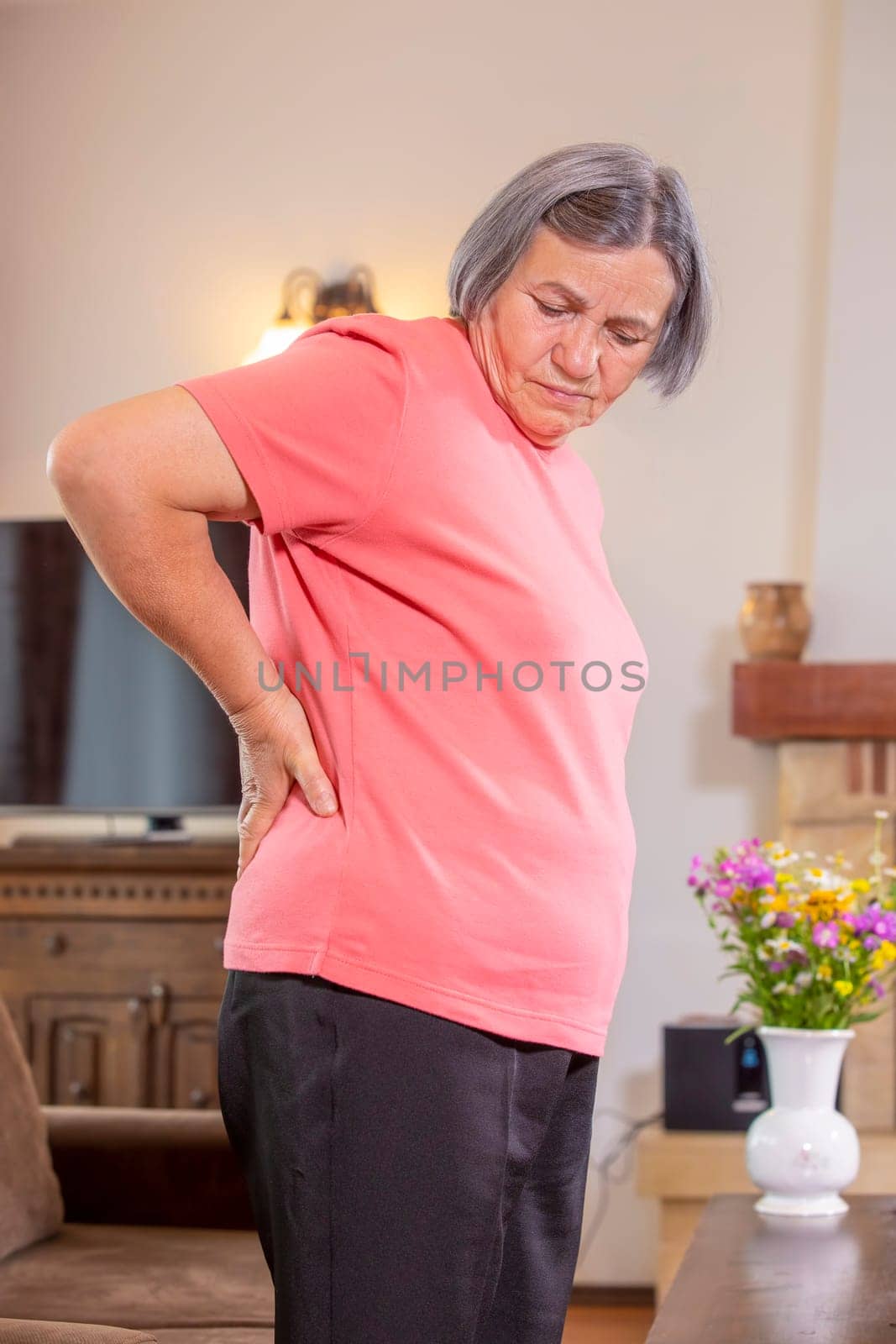 Mature woman suffering from backache at home. Massaging lower back with hand, feeling exhausted, standing in living room.