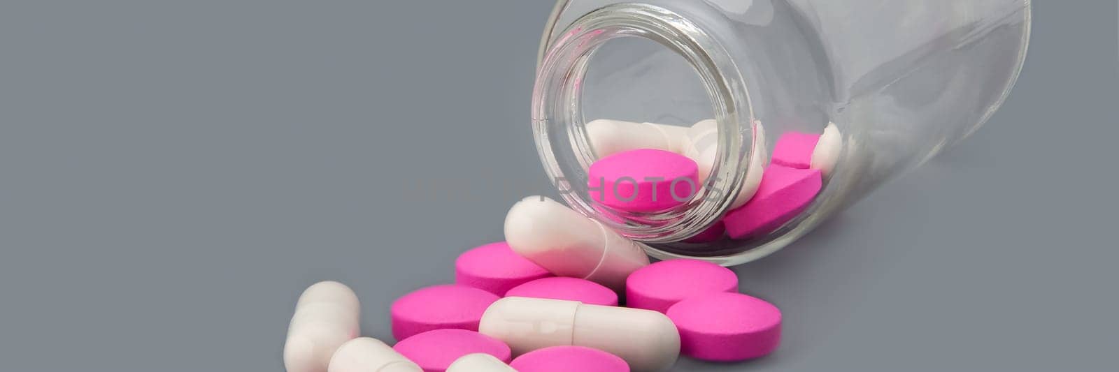 White pills and pink tablets are scattered from a glass bottle on a gray background.