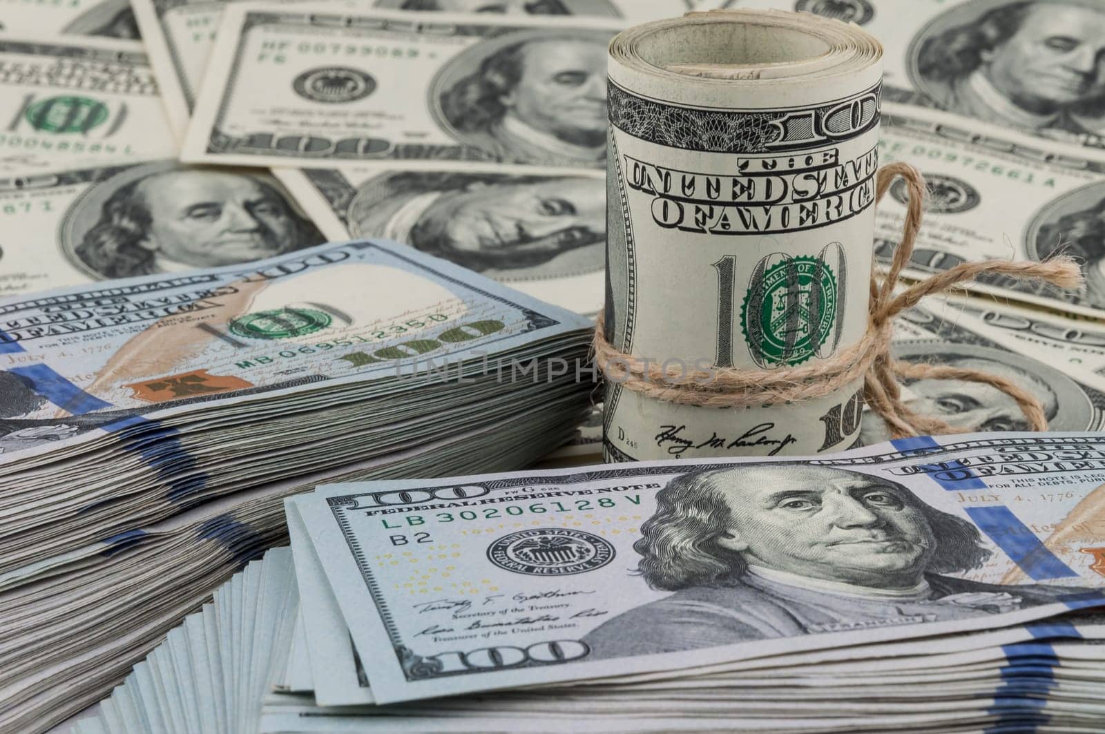 A roll of one hundred dollar bills tied with a rope against the backdrop of scattered dollars. Side view.