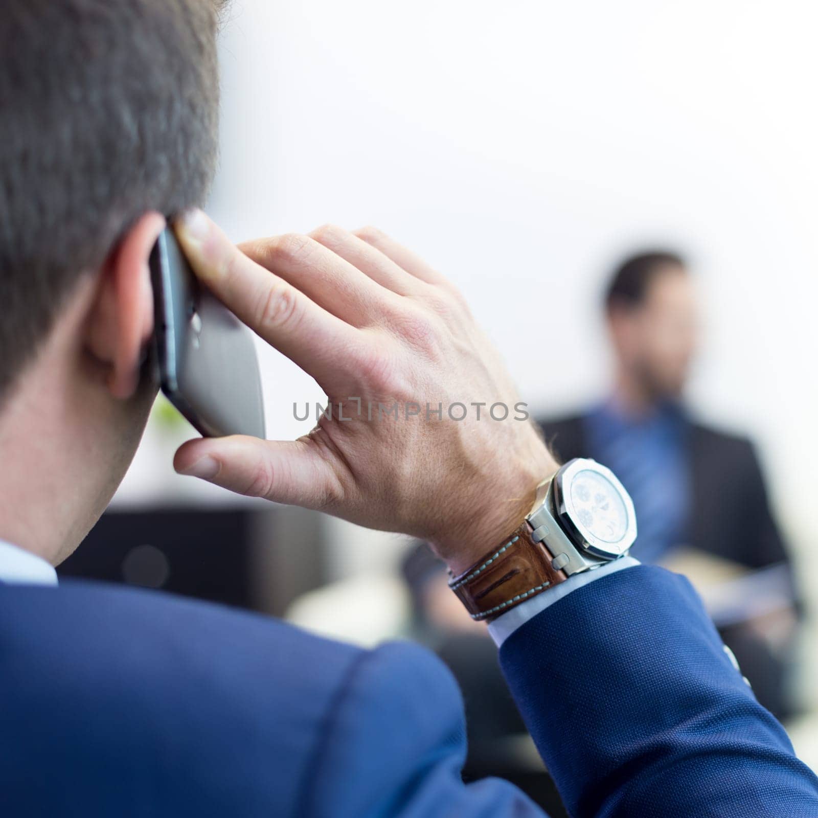 Closeup rear view of successful businessman in a stylish modern navy blue suit holding mobile phone and talking during corporate meeting. Business and entrepreneurship concept.