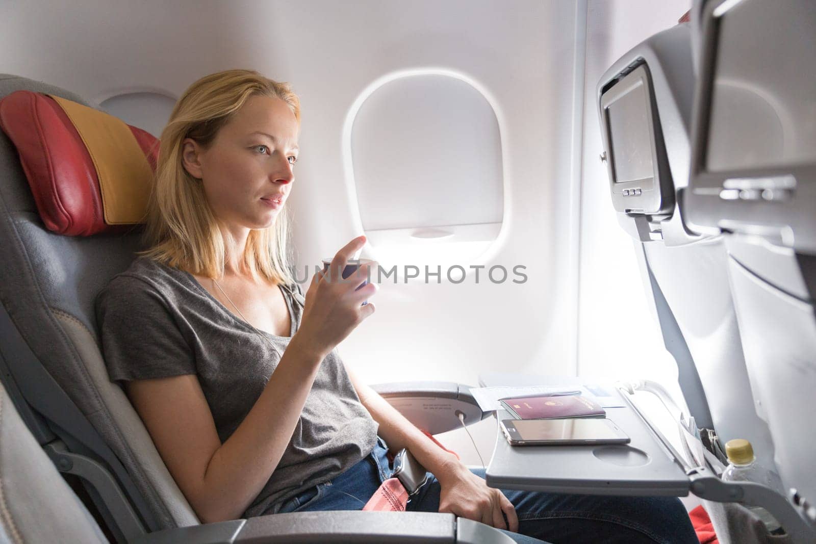 Woman on commercial passengers airplane during flight. Female traveler seated in passanger cabin drinking coffee. Sun shining trough airplane window.