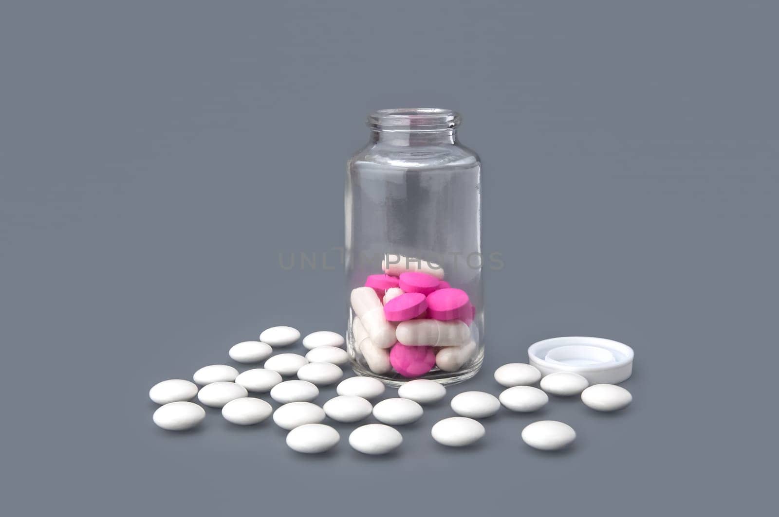 White tablets are scattered around a glass bottle with pink tablets on a gray background.