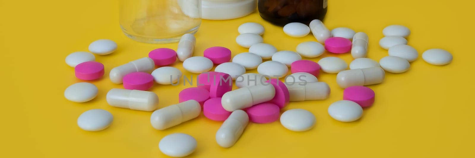 On a yellow background white and pink tablets are scattered and there are three bottles of glass and plastic. Side view.