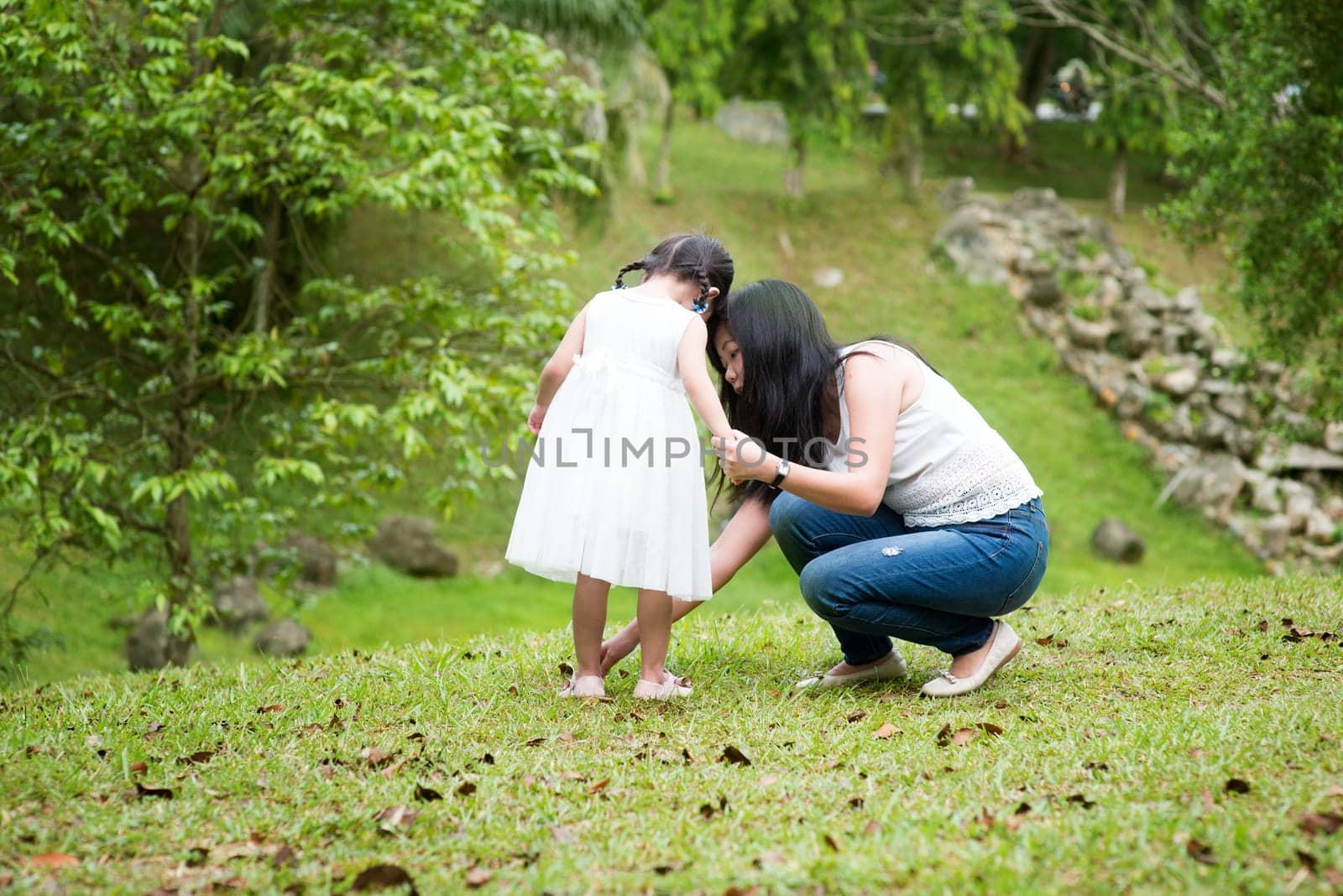Mother helps little girl wearing shoe at park. Asian family outdoors portrait.