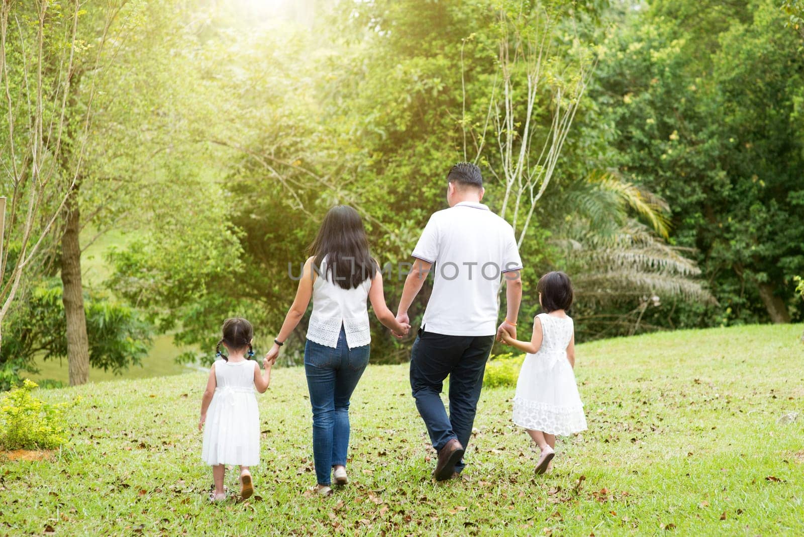 Asian family outdoors. Parents and children holding hands and walking at green park. Rear view.