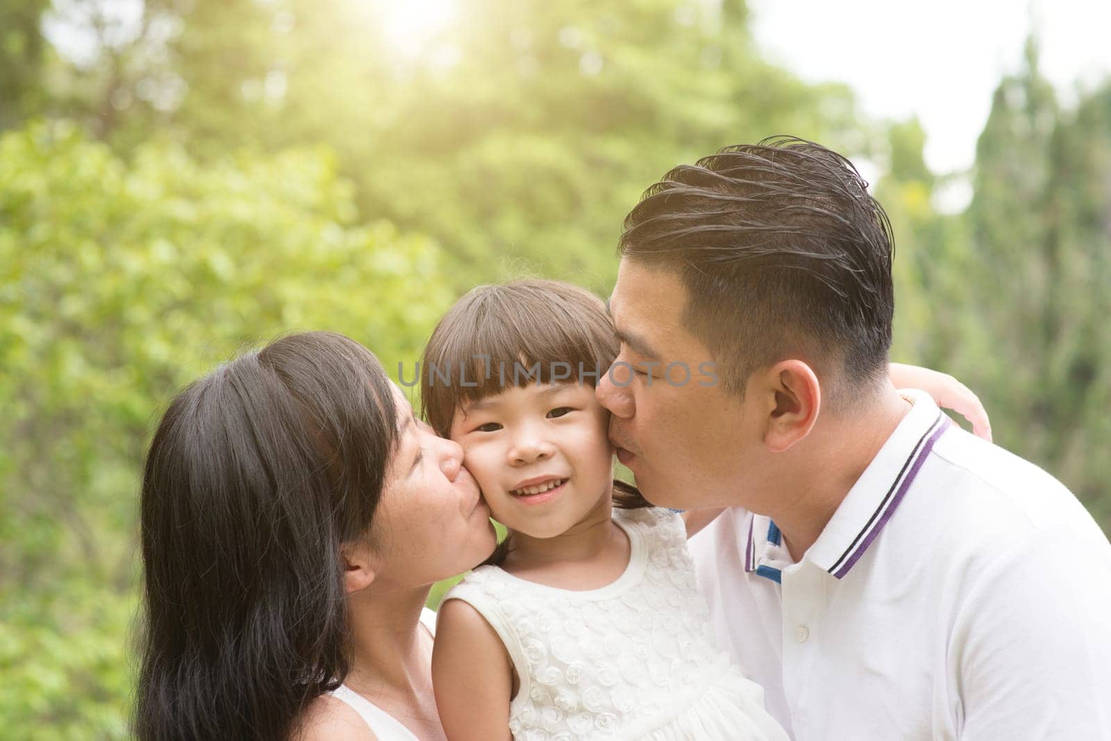 Parents kissing daughter at green park. Asian family outdoors activity.