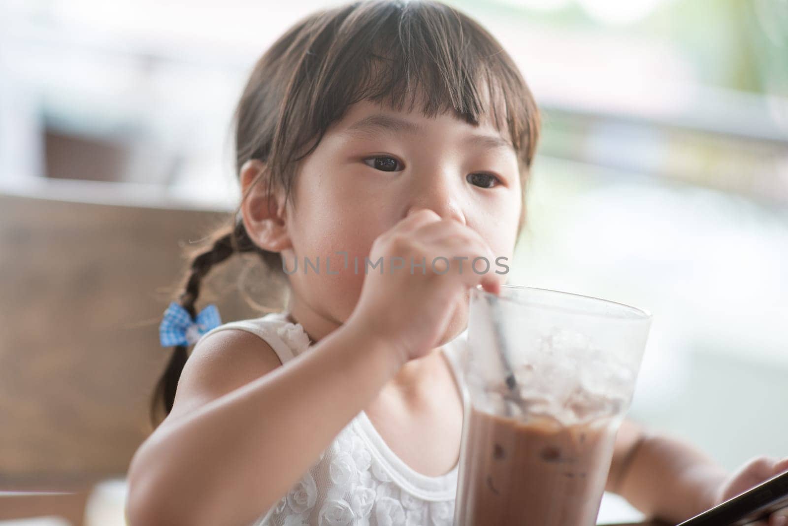 Little Asian girl drinking iced chocolate at cafe. Natural light outdoor lifestyle.