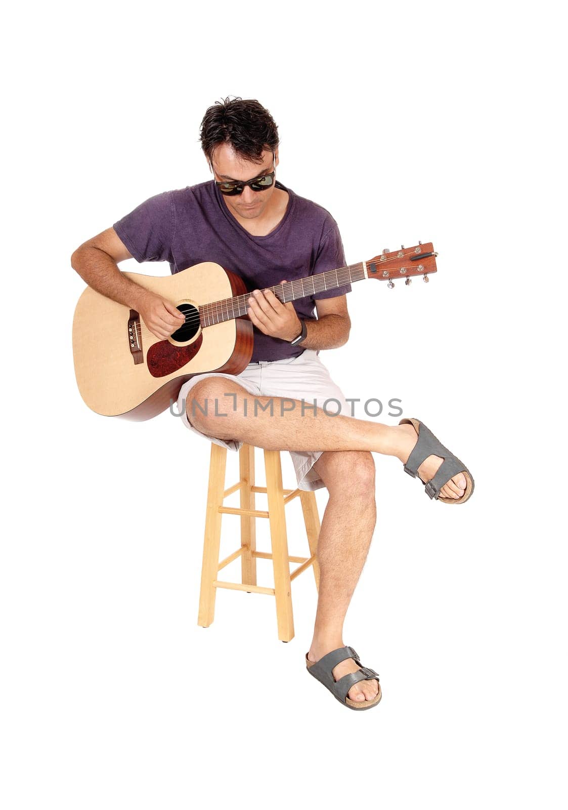A young man sitting in jeans and sunglasses, playing his guitar, looking.down, isolated for white background.