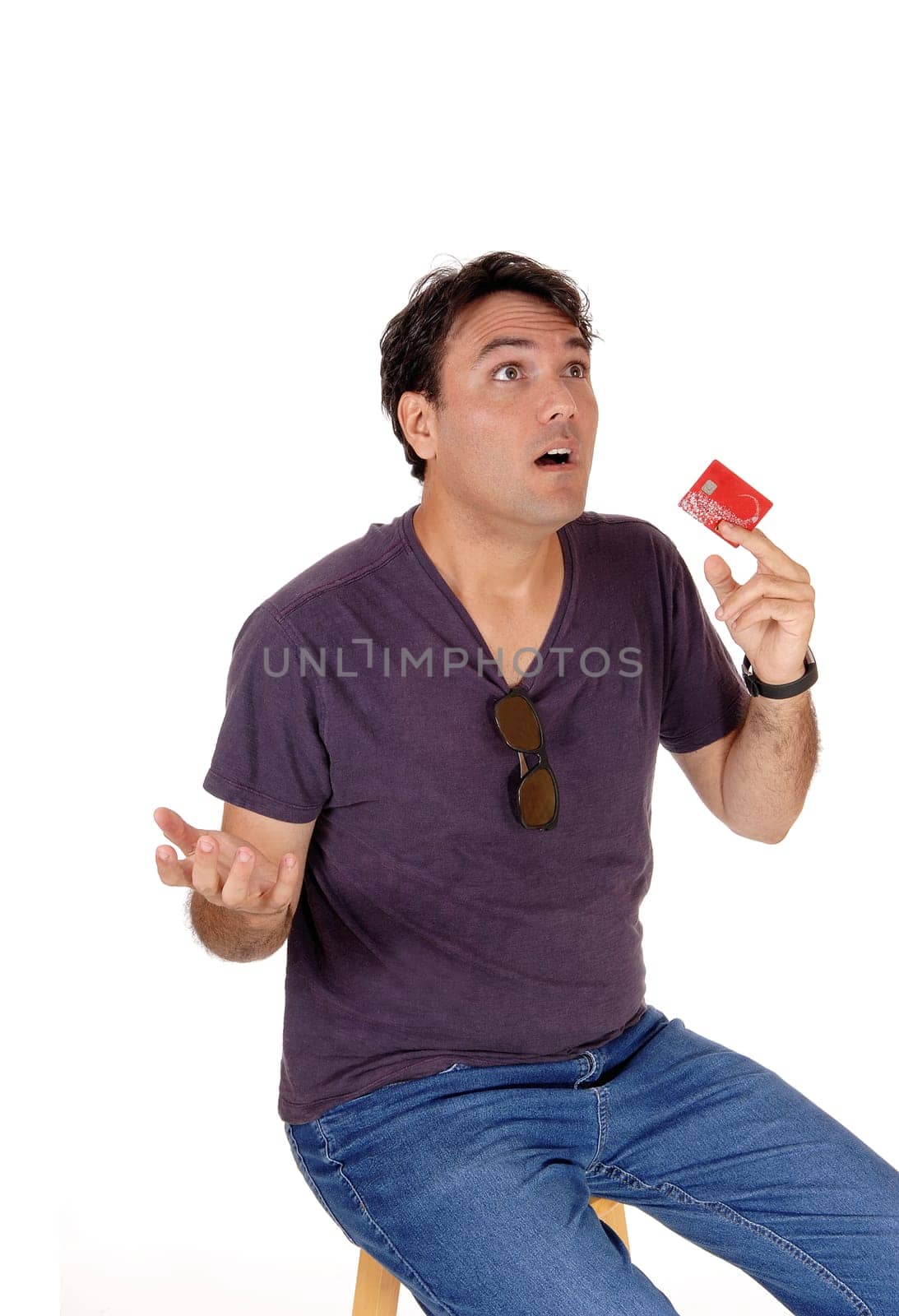 A very upset and surprised young man sitting on a chair and holding up.his credit card, spend to much, isolated for white background.