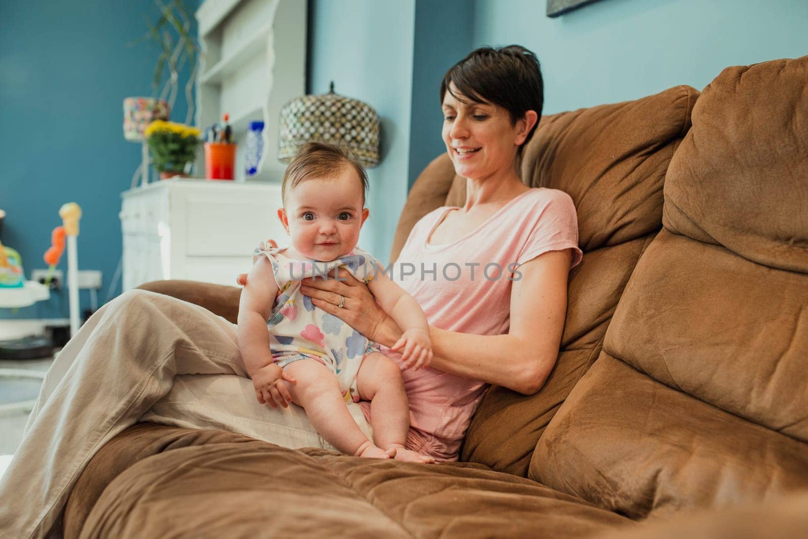 Mid adult mother sat on a living room sofa. The baby is sat up on the mothers lap being winded after having a bottle. The baby is looking at the camera and the mother is smiling at the baby.