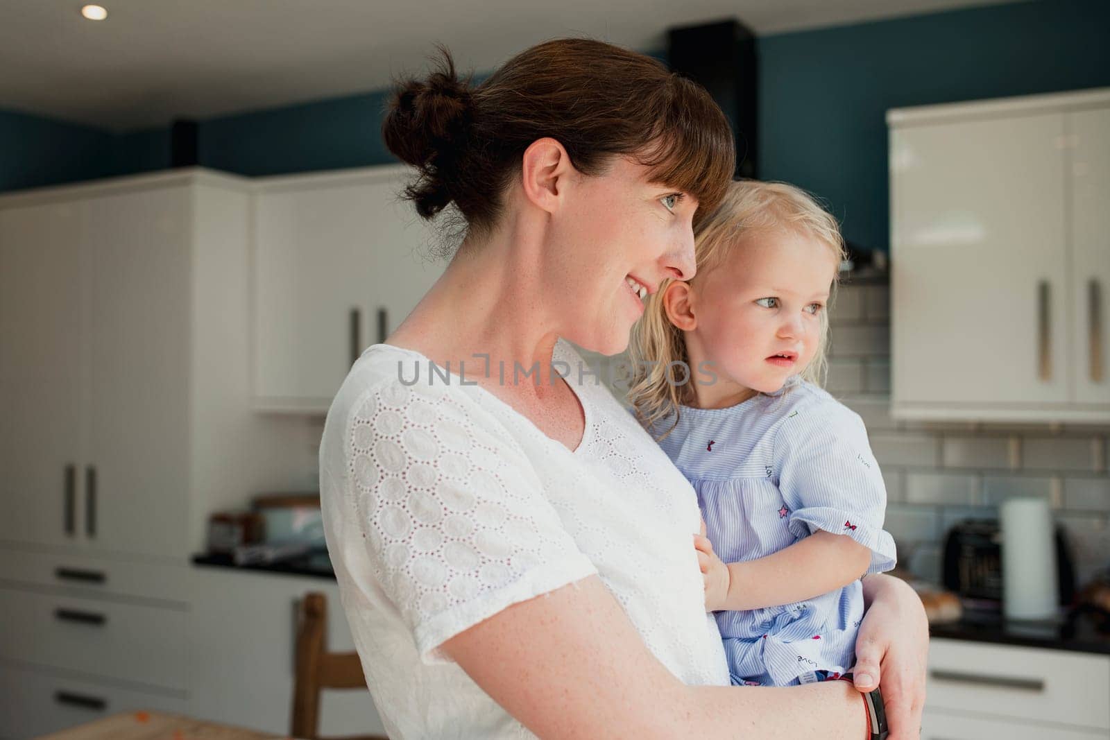 Side view of a mother holding her daughter. They are both looking out the window together while stood in the kitchen at home.