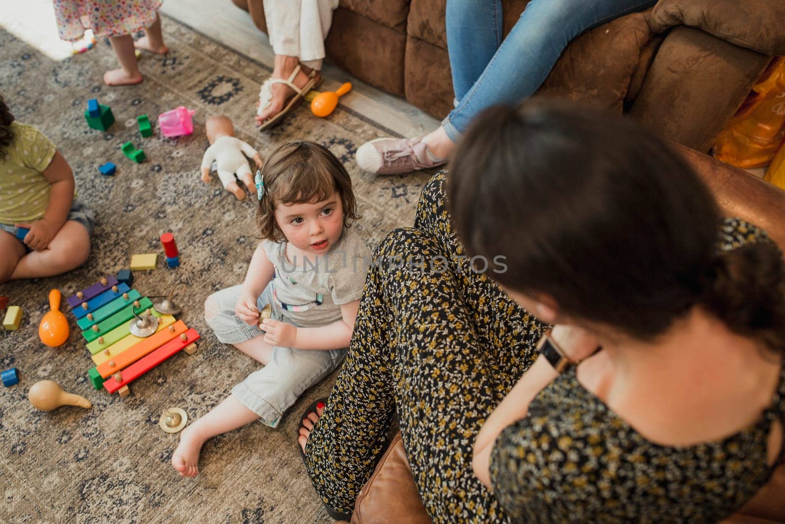High angle view of a mother looking down onto her daughter who is sat on the floor. The little girl is sat with her friend playing with toys.