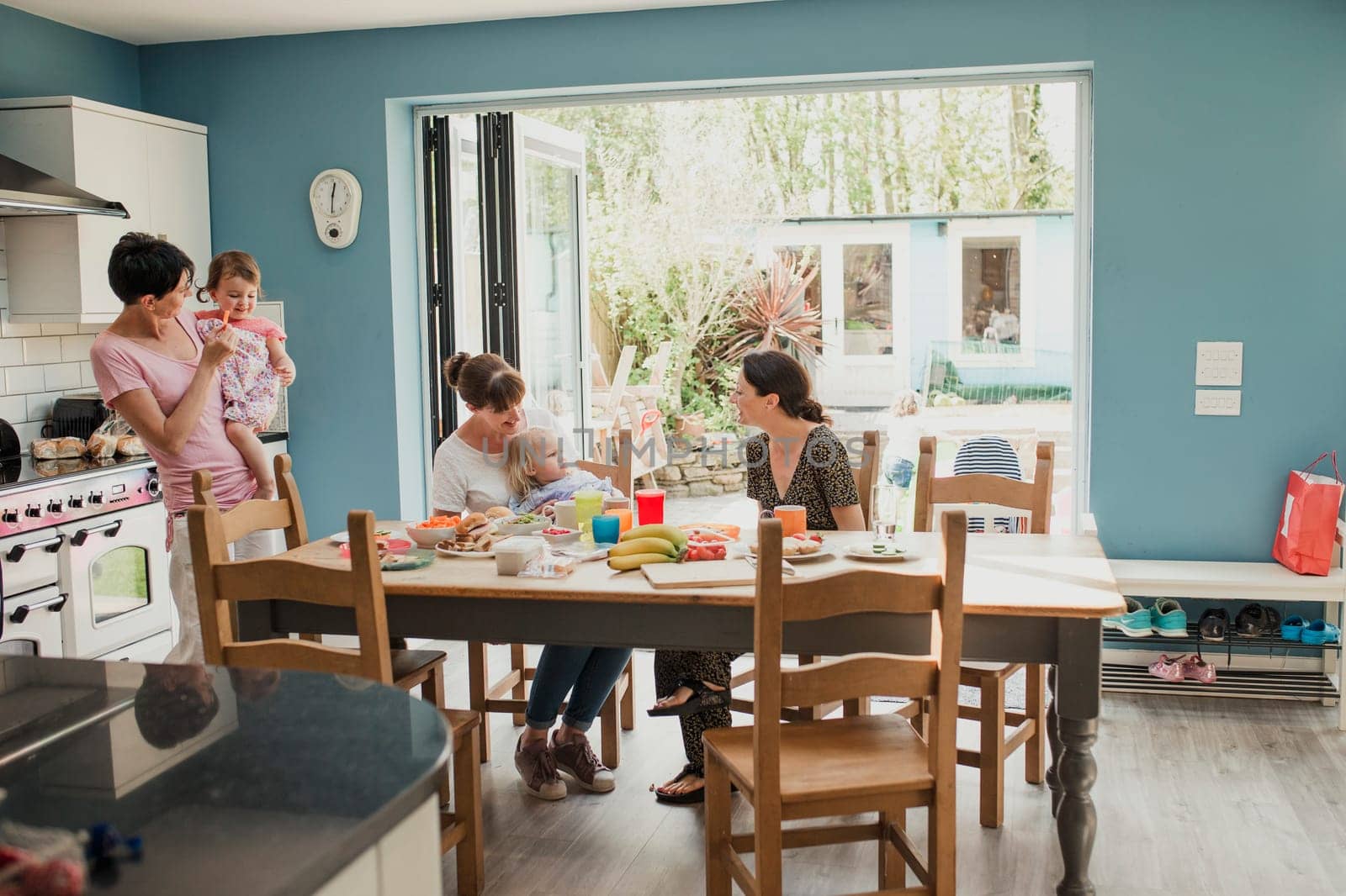 Three mid adult women standing in a kitchen at home. Two of the women are holding their children and having snacks for lunch.