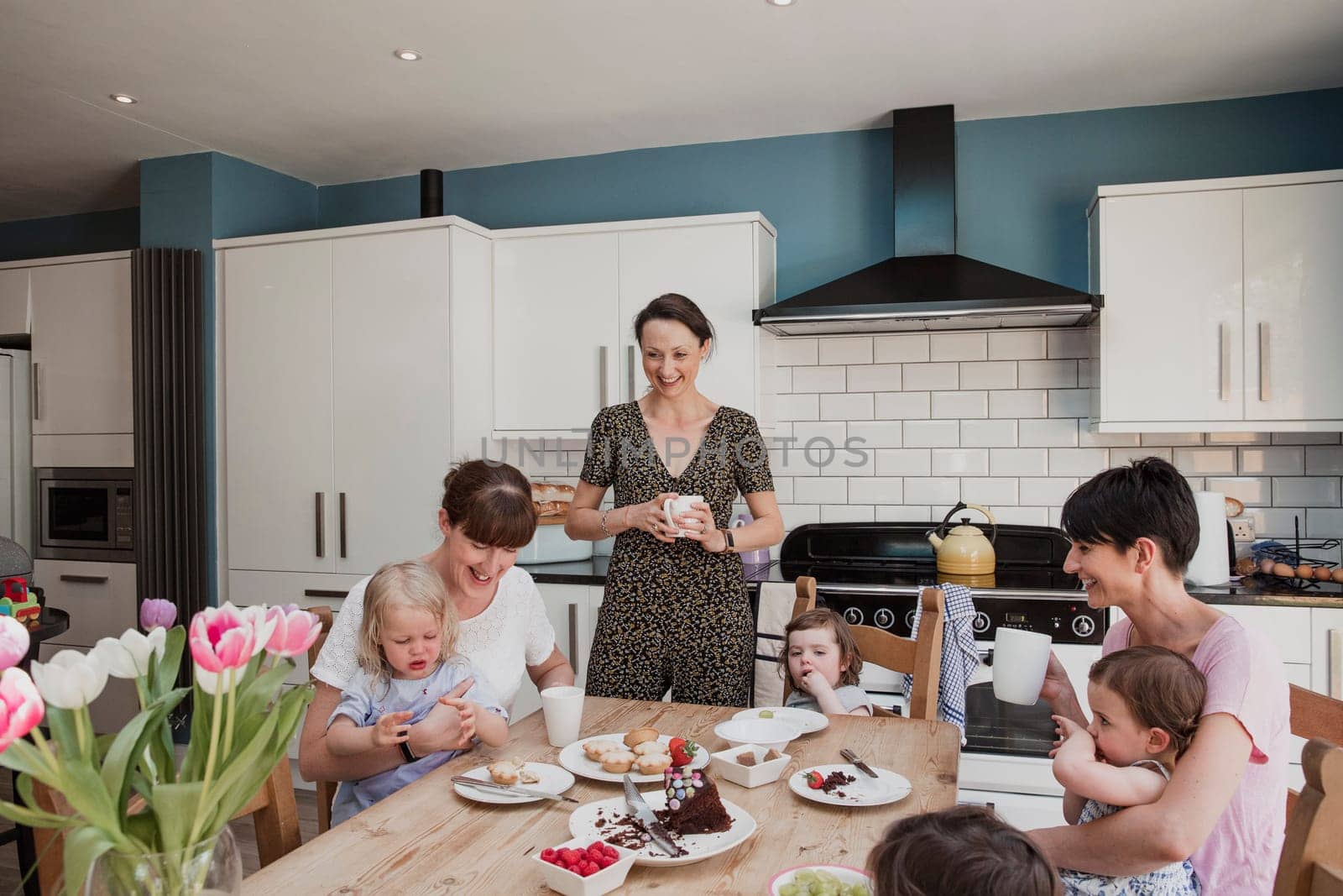 Three mid adult women in a kitchen with their daughters. Two of the women are sitting down with their daughters sitting on their laps while they eat their lunch. One mid adult woman is standing with a cup of tea and smiling.
