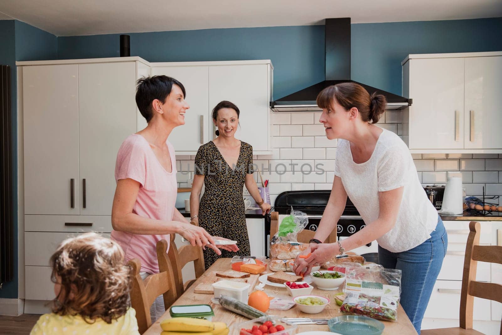 Thre mid adult women standing in the kicthen around a dining table. The women ae preparing the food for lunch. Preping some salad and sandwiches.