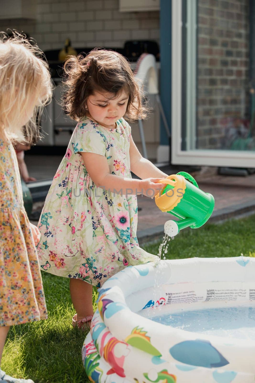 Side view of two little girls standing next to an inflateable pool. One little girl is using a toy watering can to pour water into the pool.