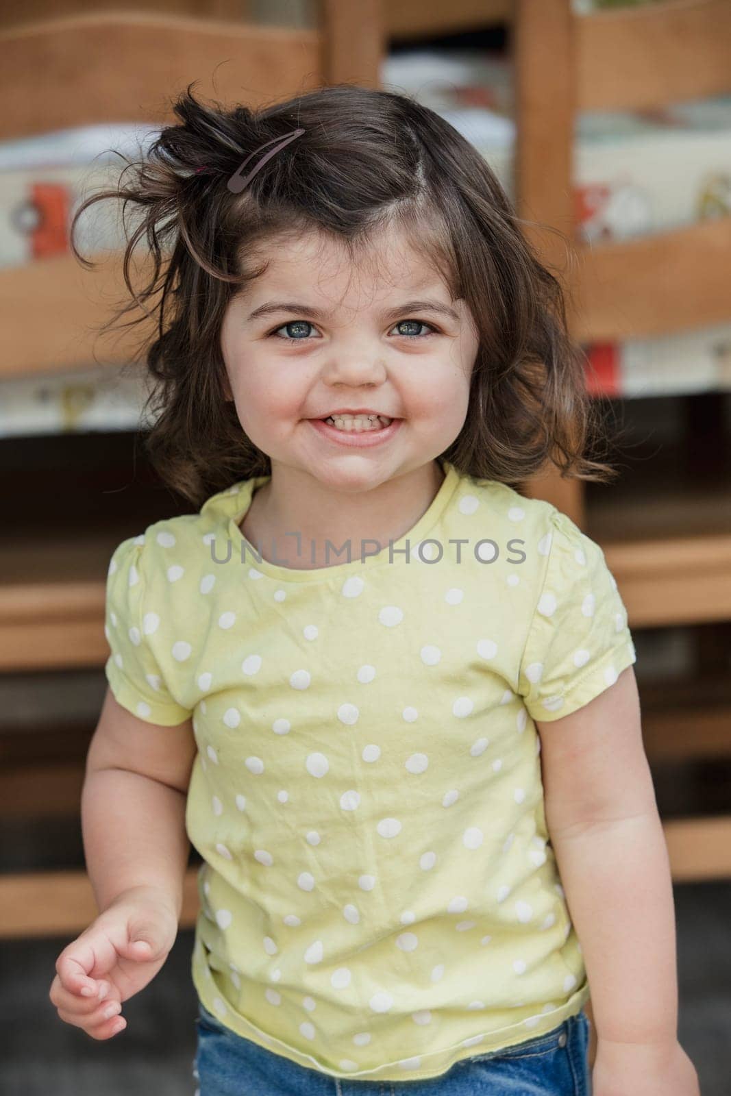 Front view of a little girl standing indoors, looking at the camera and smiling for a headshot. The little girl is wearing casual clothing.