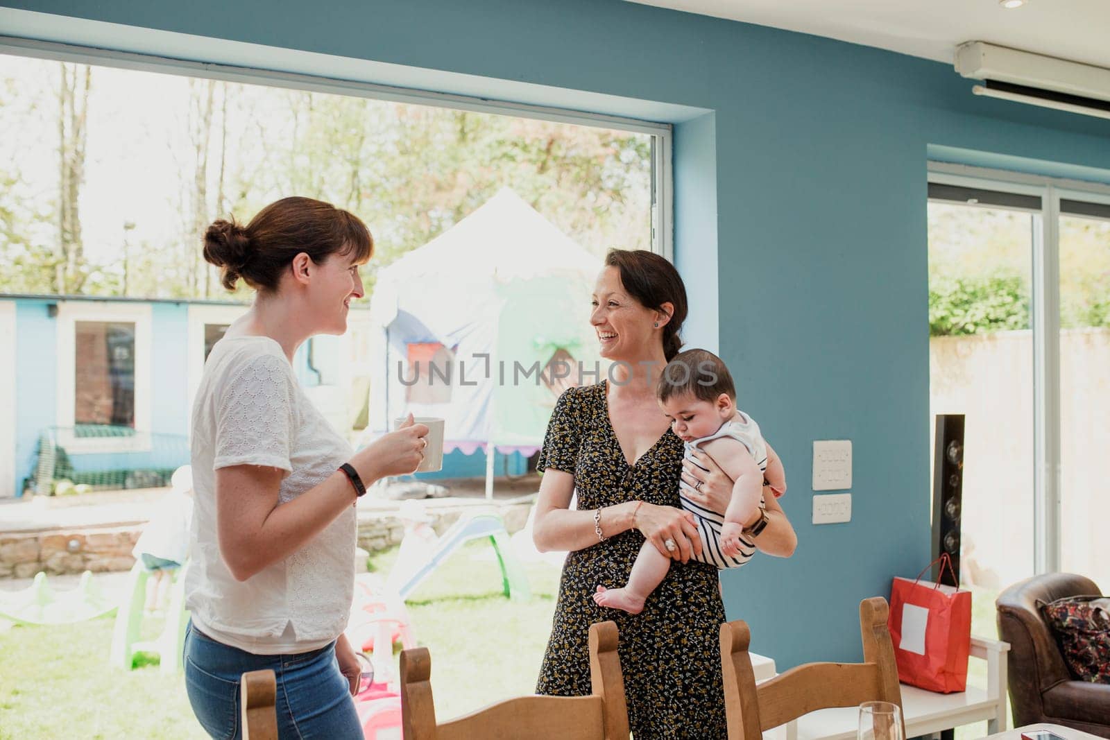 Two mid adult women standing in the kicthen at home and socialising. One woman is holding her baby son. They are talking and laughing together.
