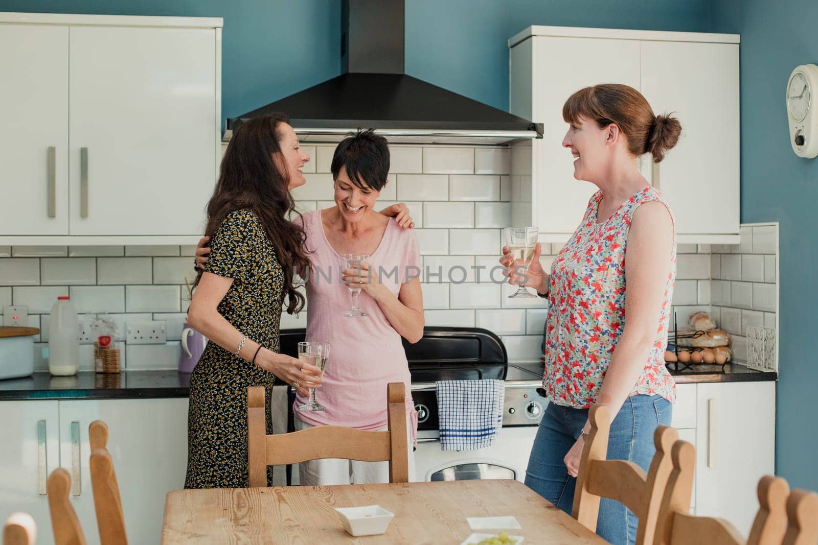Three mid adult women standing in a kicthen enjoying a glass of wine. The women are all standing around the stove in the kitchen, talking and laughing.