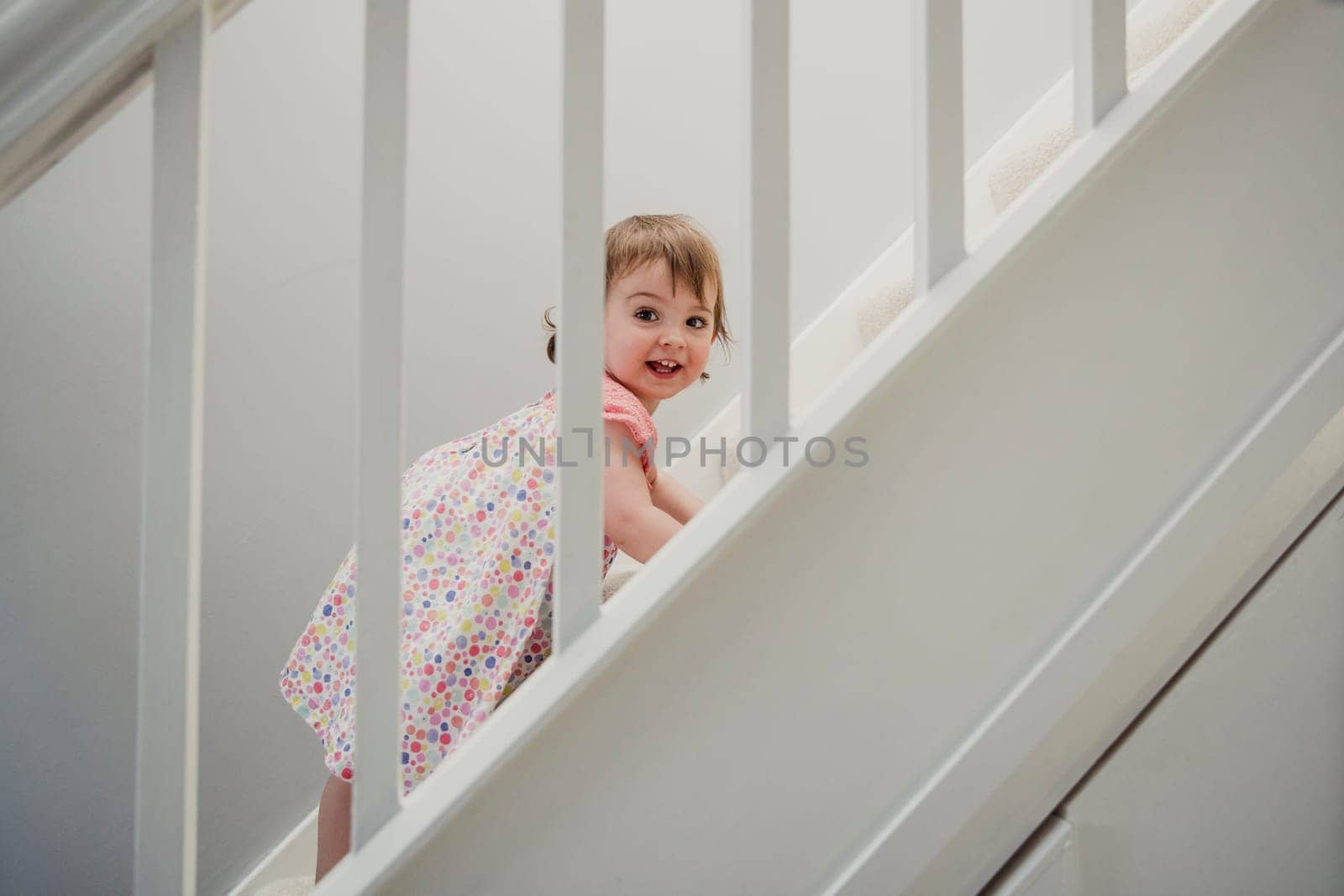 Side view of a staircase in a house. There is a toddler girl climbing up the stairs. She is turning her head and smiling while looking at the camera.