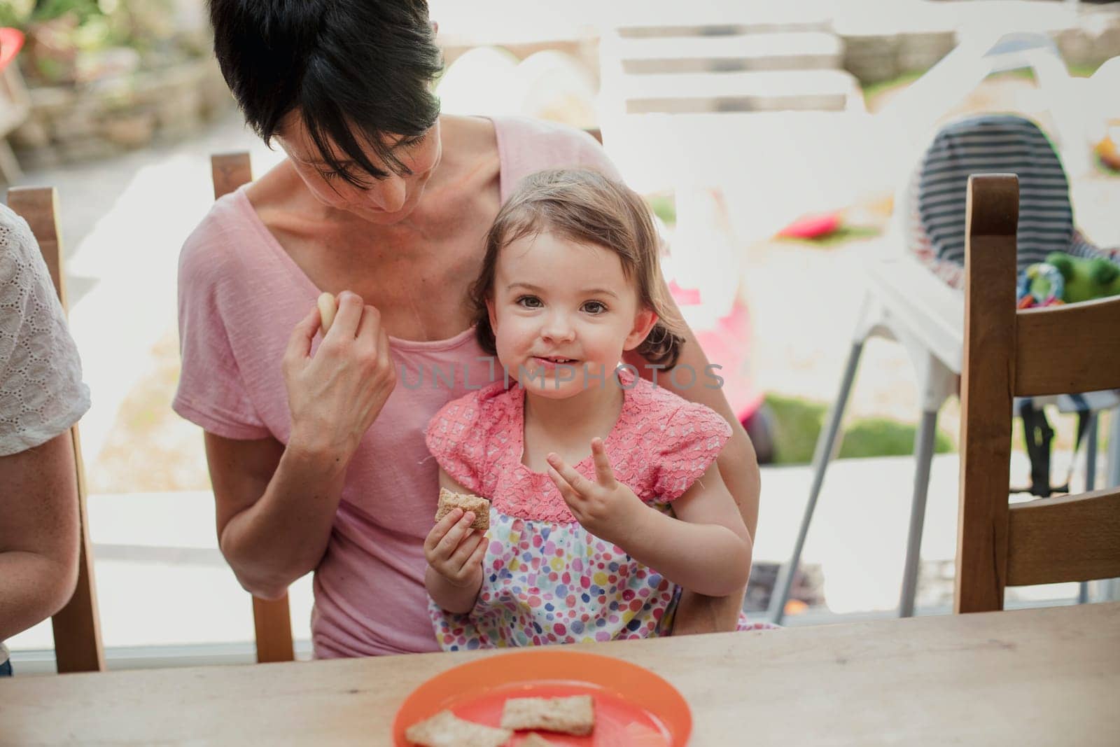 Front view of a little girl looking at the camer and enjoying a little sandwich. The little girl is sitting on her mothers lap, her mother is looking down at her and waiting to give her some more food.