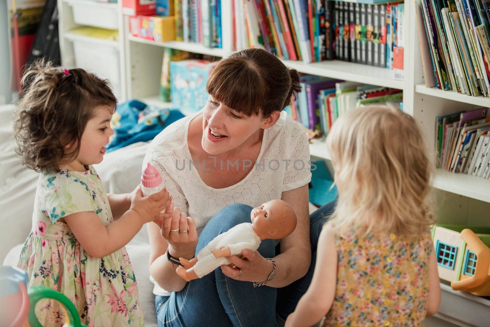 Mother and her two daughters are looking at a toy baby doll and talking about giving it a bottle. The mother is holding the doll baby and passing her daughter the toy baby bottle.