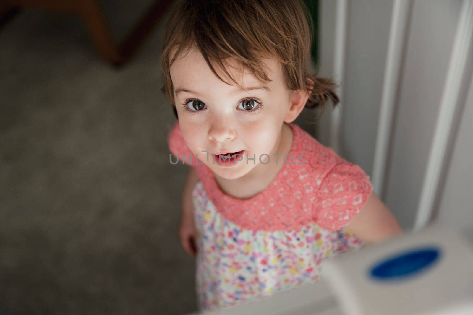 HIgh angle view of a little girl standing on the other side of a saftey baby gate. The little girl is looking up at the camera with a cheeky little smile.