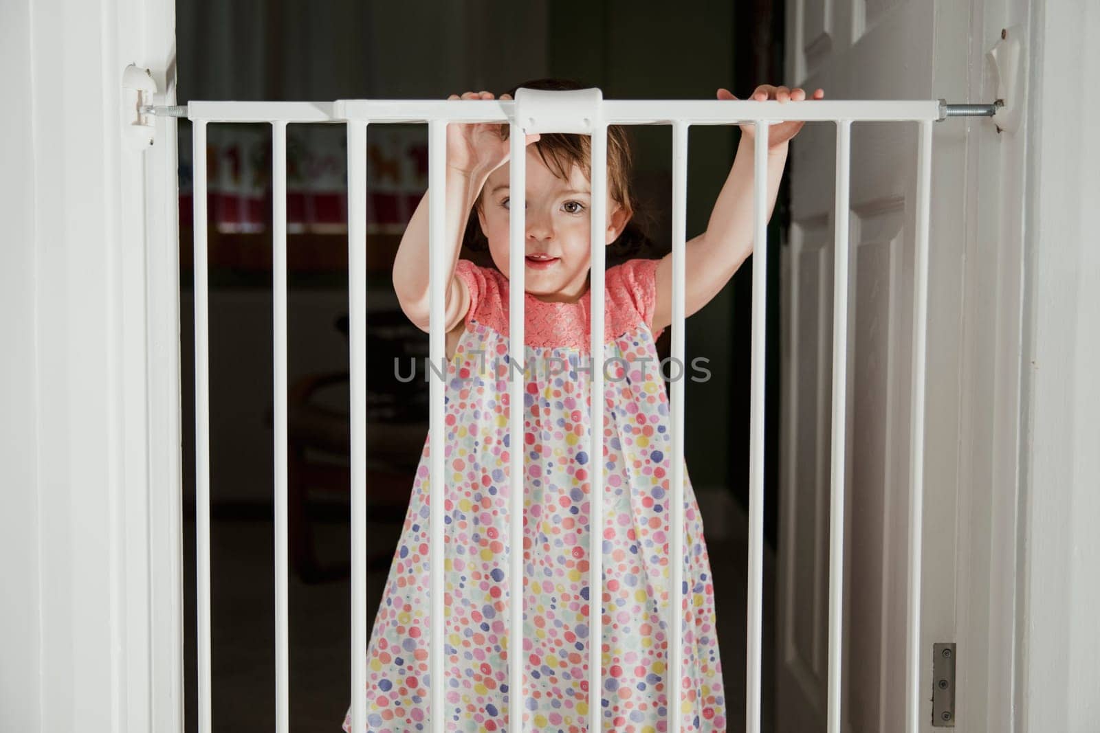Front view of a little girl standing on the other side of a baby saftey gate. The little girl is holding onto the top of the saftey gate and looking at the camera over the top.