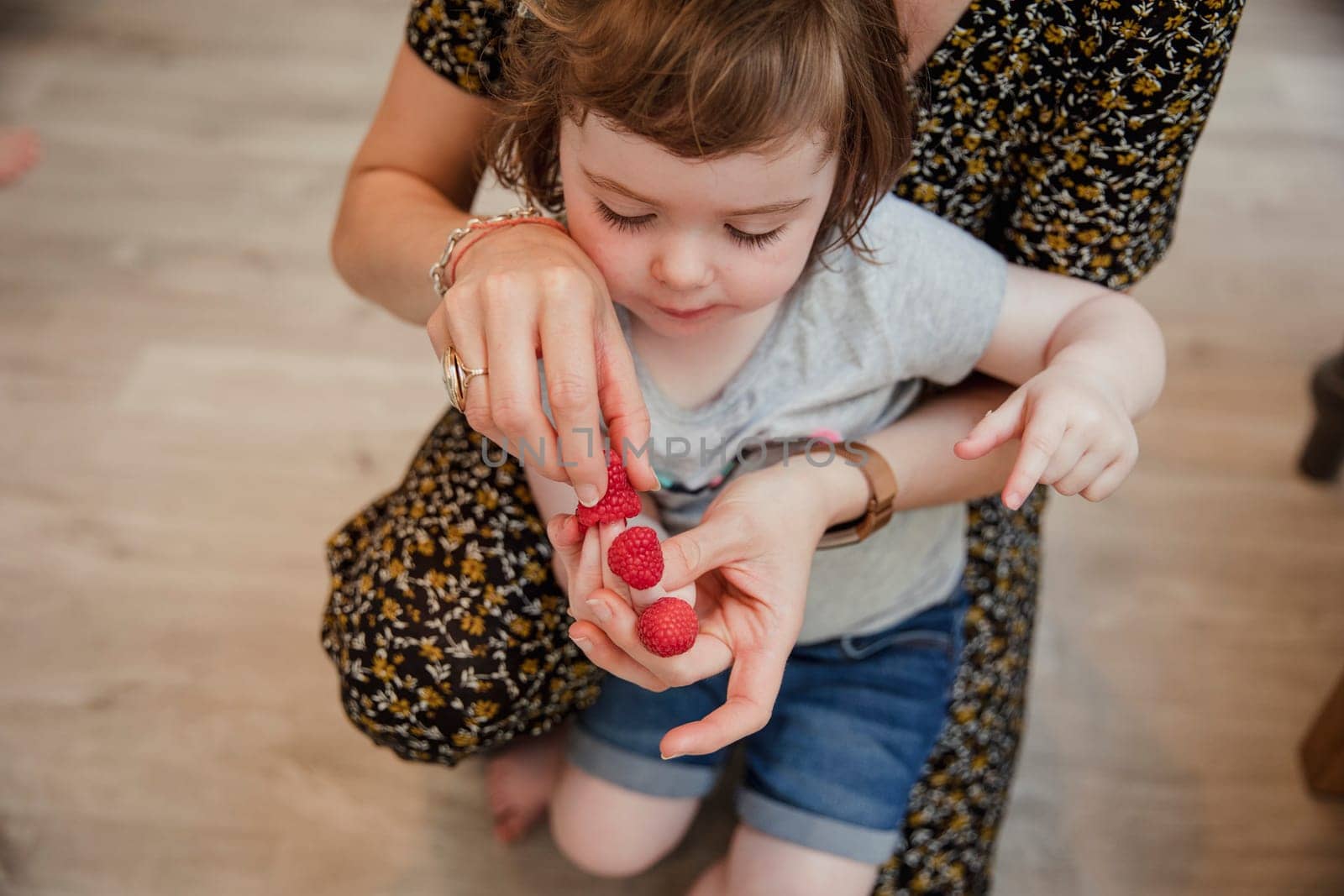 Front view of a little girl playing with some raspberries and putting them onto her fingers. Her mother is kneeling behind her helping and laughing with her.