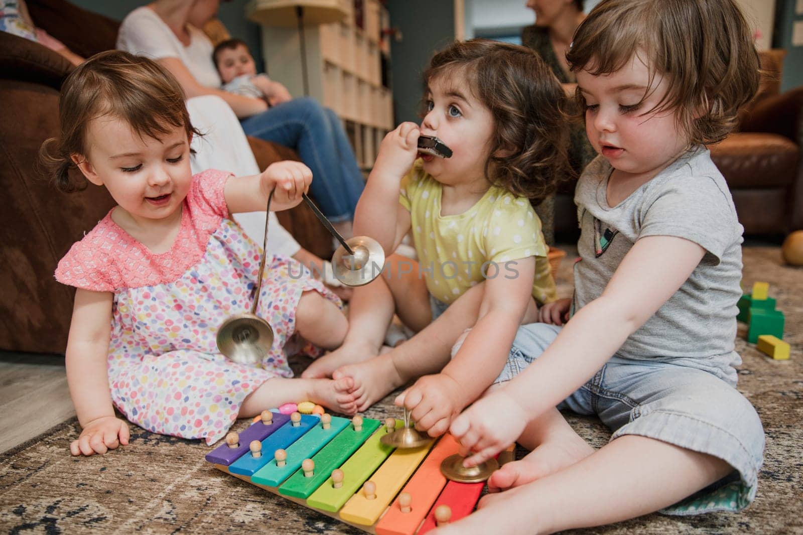 Three little girls sitting in on the floor in a living room playing together and having fun. They are each playing different musical instruments  symbols, xylophone and harmonica.