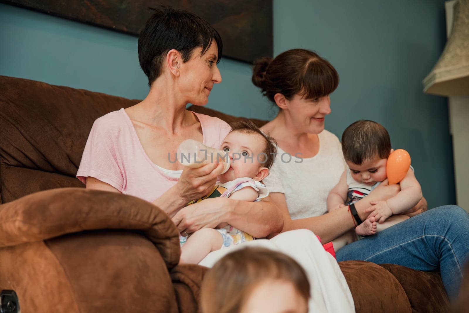 Mid adult mother is feeding her baby daughter a bottle of milk. sitting next to her is her mid adult friend with her baby boy sitting on her lap and winding him.