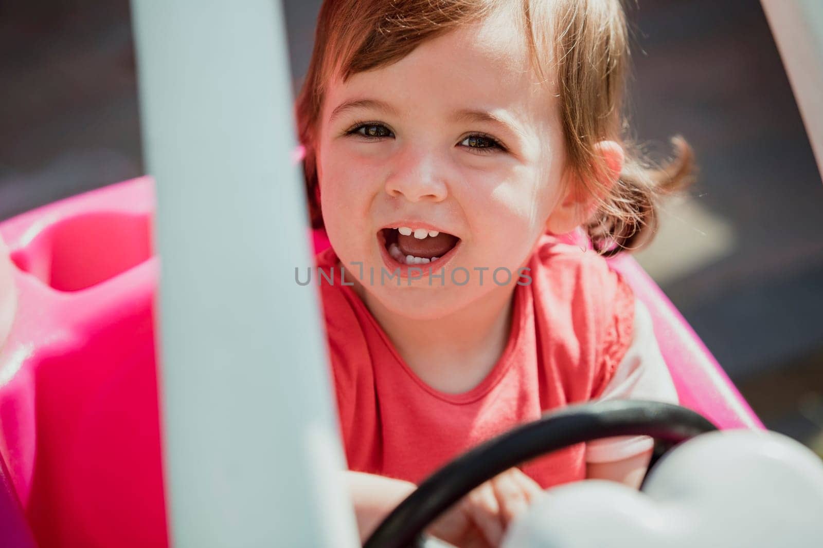 Little girl looking up at the camera and smiling with her mouth open. She is sat in a toy car, excited to play and have fun.