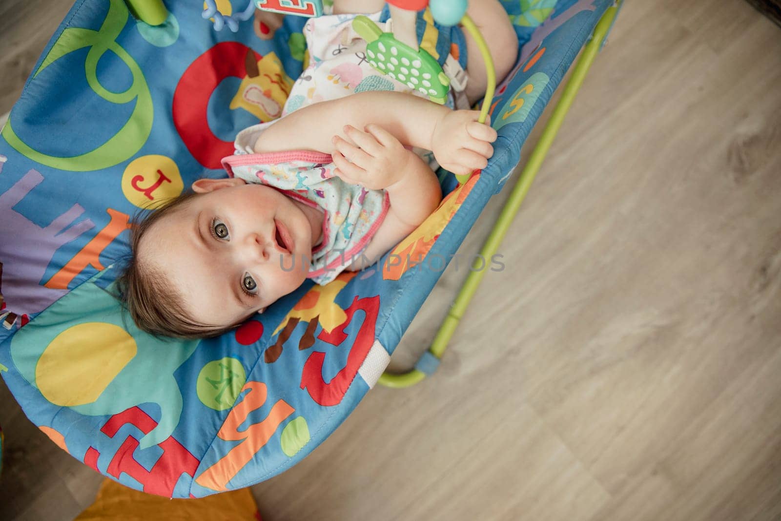 High angle view looking down onto a baby girl relaxing in her baby bouncer. The baby is looking up into the camera and smiling.