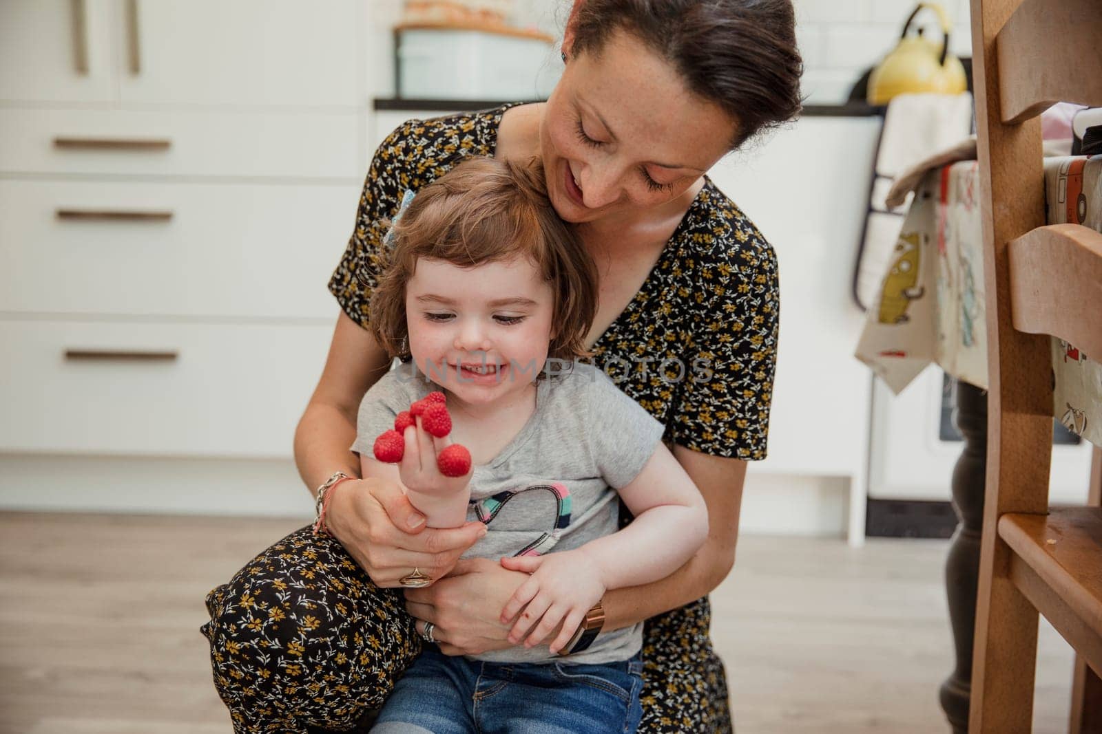 Front view of a little girl playing with some raspberries and putting them onto her fingers. Her mother is kneeling behind her helping and laughing with her.