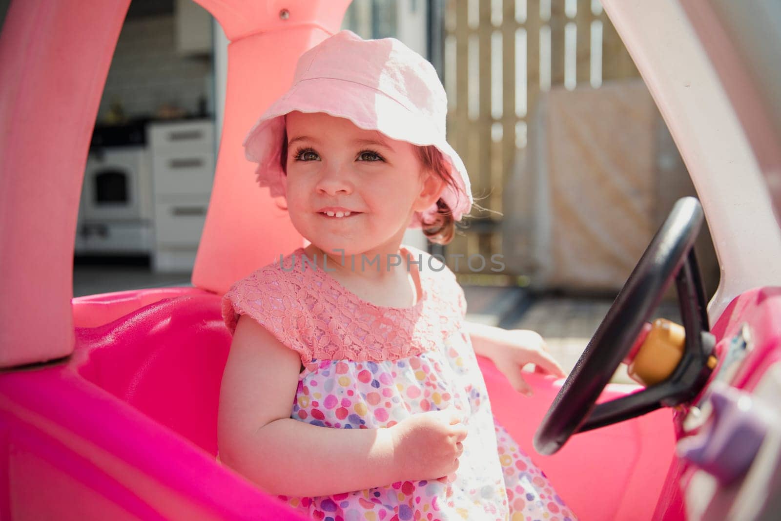 One little girl sitting outside in her toy car using her imagination. She is looking out of the window, smiling and enjoying herself.