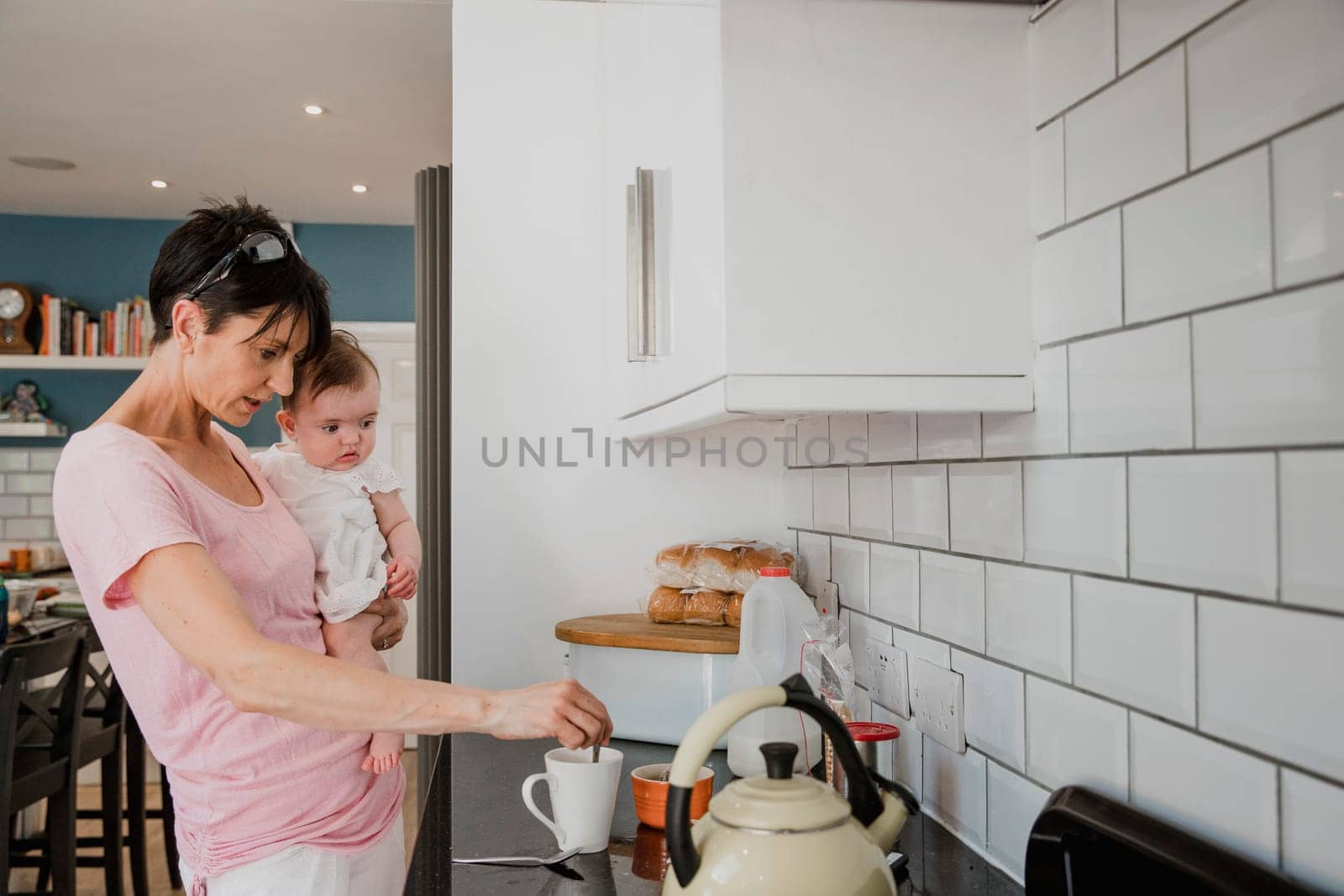 Side view of a mother holding her baby daughter while standing in the kitchen. The woman is making a cup of tea while carrying her daughter on her side.