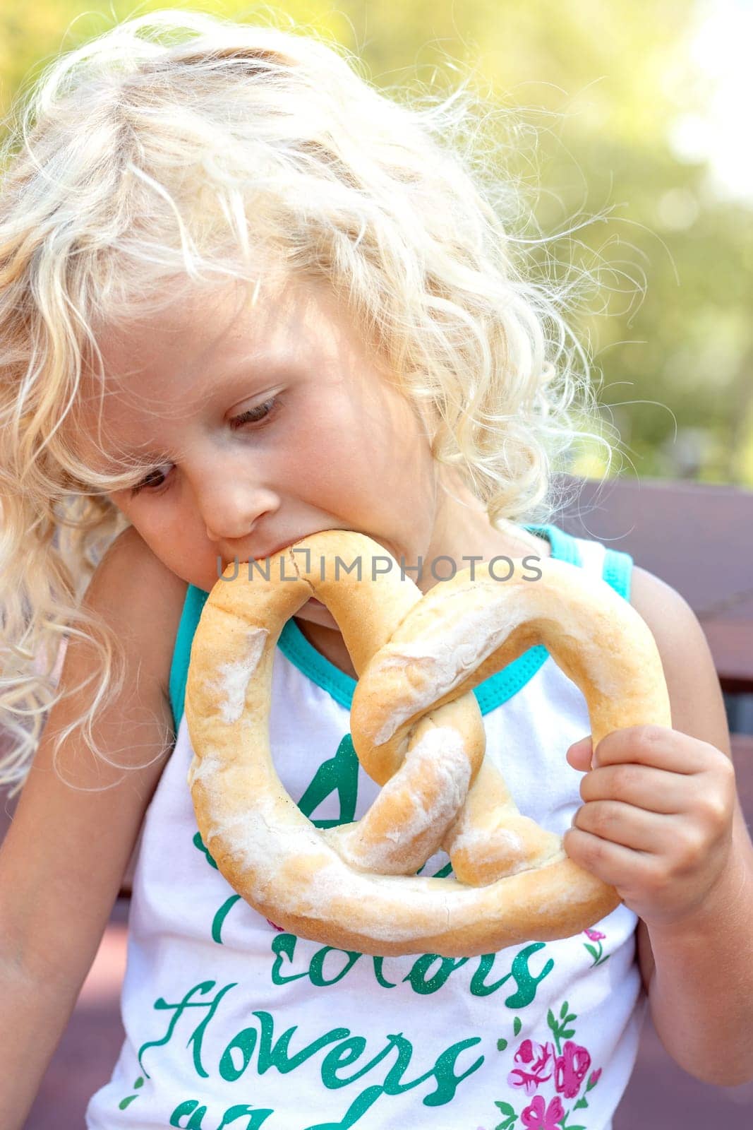 Adorable little girl sitting on the bench with her pretsel in park. Blonde small girl eating outdoors.
