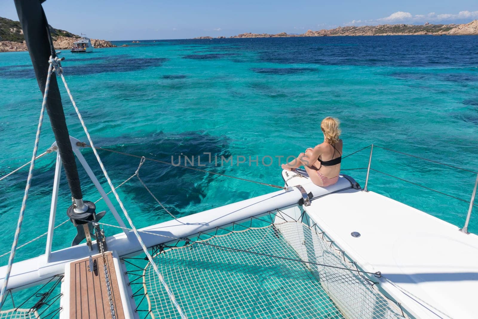 Woman in bikini tanning and relaxing on a summer sailin cruise, sitting on a luxury catamaran in picture perfect turquoise blue lagoon near Spargi island in Maddalena Archipelago, Sardinia, Italy.