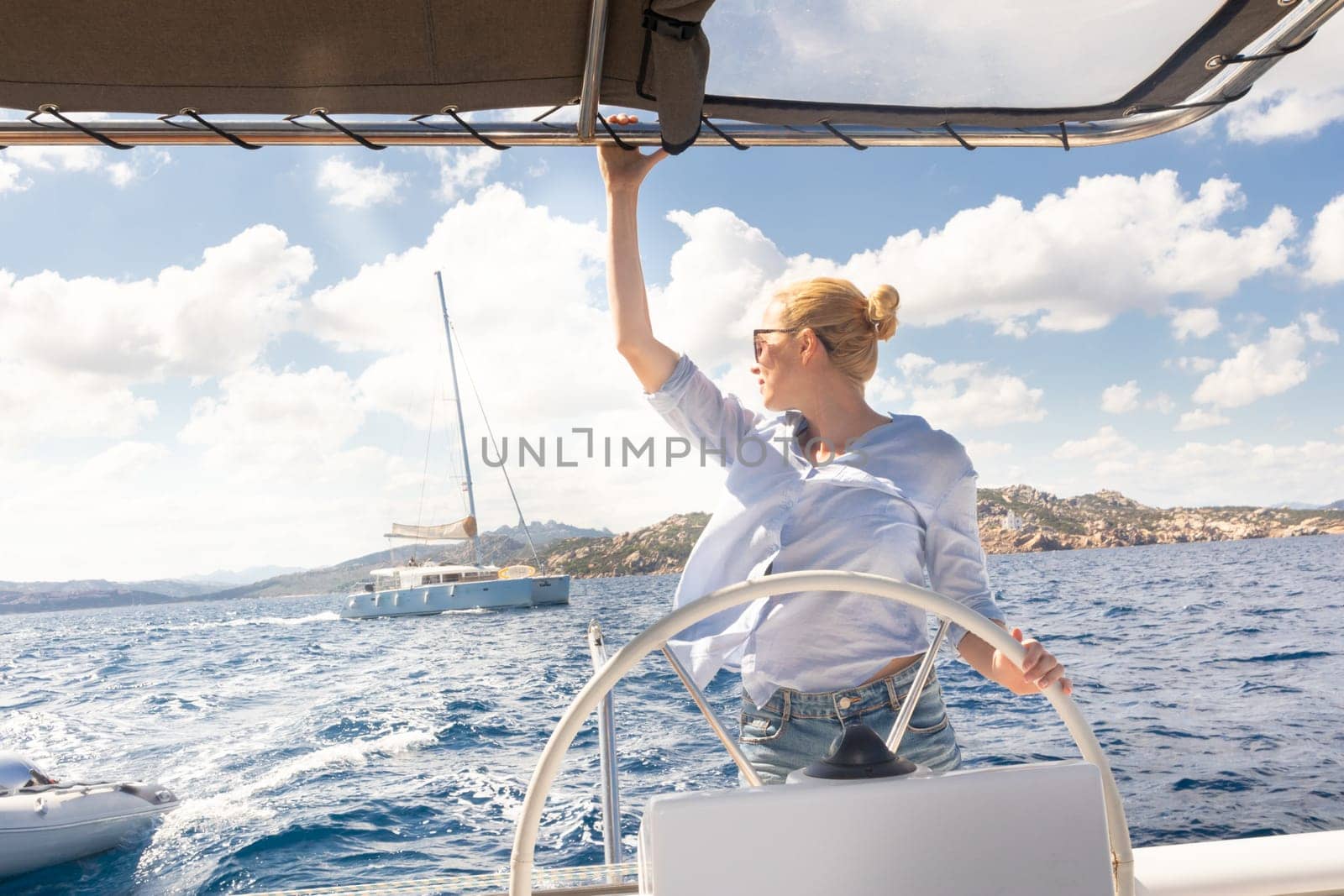 Attractive blond female skipper navigating the fancy catamaran sailboat on sunny summer day on calm blue sea water. Luxury summer adventure, active nautical vacation.
